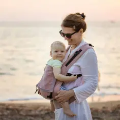 Uśmiechnięta mama z dzieckiem w chuście na plaży. Dziecko wygląda jakby miało wieniec laurowy na głowę, otoczone miłością.