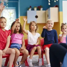 Dzieci na zajęciach w przedszkolu, z podniesionymi rękami. Widać, ile lat dziecko chodzi do przedszkola, bo są w różnym wieku.