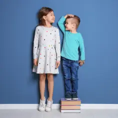 Dzieci mierzą wzrost, zastanawiając się, ile cm rocznie rośnie dziecko. Chłopiec stoi na książkach, by dorównać dziewczynce.