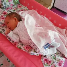 Nowe dziecko śpi otulone w pieluchę tetrową na różowej macie. Pokazano, jak zawijać dziecko w pieluchę tetrową, by czuło się bezpiecznie.