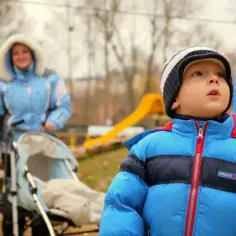Chłopiec w niebieskiej kurtce na placu zabaw, obok wózek i kobieta. Może to być moment przed tym, jak dowiedzieć się, jak wypisać dziecko z przedszkola.