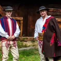 Dwóch młodych mężczyzn w tradycyjnych strojach ludowych. Jeden ma na sobie kamizelkę, drugi narzucił na ramię brązowy płaszcz. Strój ludowy dla chłopca jest tu pięknie wyeksponowany.