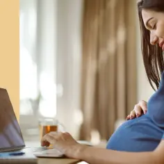 Kobieta w ciąży, uśmiechnięta, sprawdza w internecie, jak liczyć ruchy dziecka. Na biurku laptop i kubek herbaty.