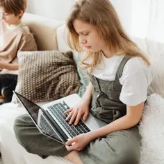 Dziewczynka na laptopie szuka informacji, jak sprawdzić lokalizację dziecka. Na kanapie obok pies i chłopiec z książką.