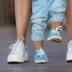 Dziecko w niebieskich bucikach stawia pierwsze kroki, a obok jego stóp widać białe trampki mamy. Widać, ile większy but dla dziecka.