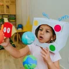 Dziecko w stroju kosmonauty z kartonowym hełmem i planetami w rękach. Wygląda jakby właśnie wyruszało w kosmiczną podróż.