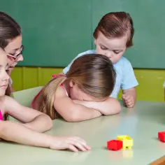 Dzieci przy stole z klockami. Jedno dziecko chowa twarz w ramionach, być może zastanawiając się, ile trwa adaptacja w przedszkolu.