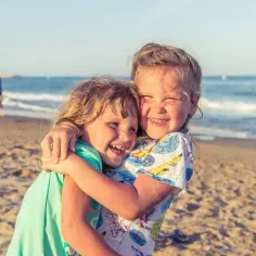 Dwie uśmiechnięte dziewczynki przytulają się na plaży. Idealne miejsce, gdzie pojechać z rocznym dzieckiem, by cieszyć się słońcem i morzem.
