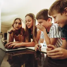 Dzieci szukają w internecie, jak usunąć blokadę rodzicielską z telefonu. Jedno z nich pisze na laptopie, inne patrzy z zaciekawieniem.