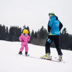 Mała dziewczynka w różowym kombinezonie i żółtym kasku uczy się jeździć na nartach z instruktorem. Idealne miejsce, gdzie na narty w Polsce z dziećmi.