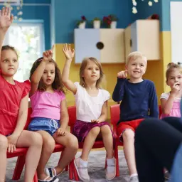 Dzieci na zajęciach w przedszkolu, z podniesionymi rękami. Widać, ile lat dziecko chodzi do przedszkola, bo są w różnym wieku.