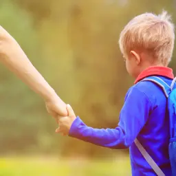 Chłopiec w niebieskim plecaku, trzymający mamę za rękę, idzie do przedszkola. Ma około 4-5 lat.