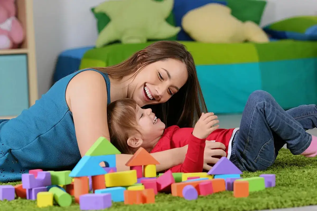 Uśmiechnięta opiekunka i dziecko bawią się klockami. Zastanawiasz się, ile kosztuje opiekunka do dziecka?