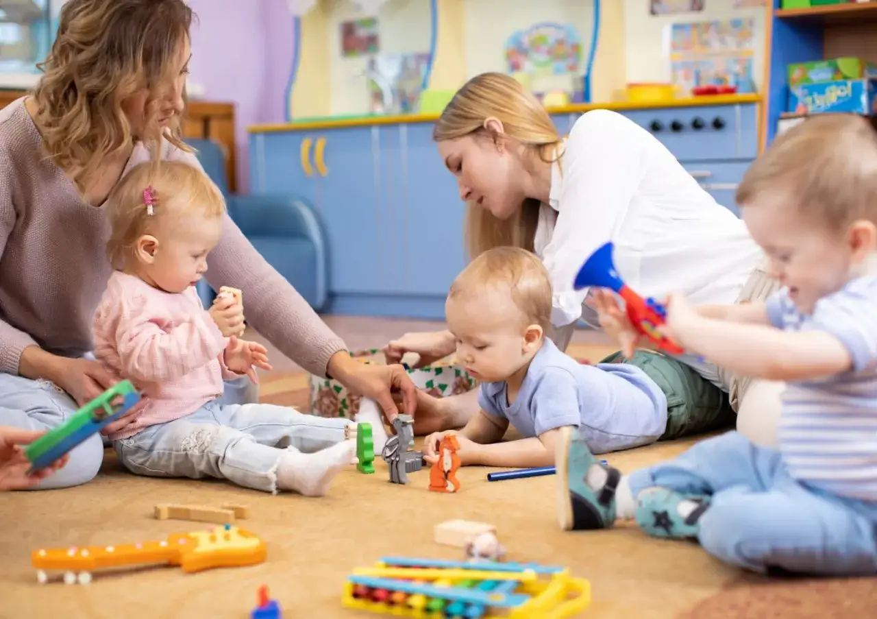 Dwie opiekunki bawią się z czwórką dzieci na dywanie. W żłobku jest 2 dzieci na opiekuna.