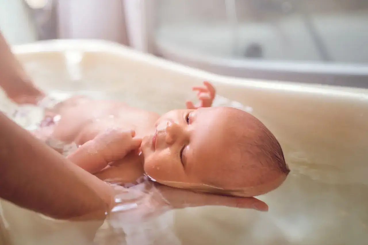 Niemowlę w wanience, delikatnie podtrzymywane przez dłoń. Czy można myć dziecko z gorączką? Kąpiel w letniej wodzie może pomóc.