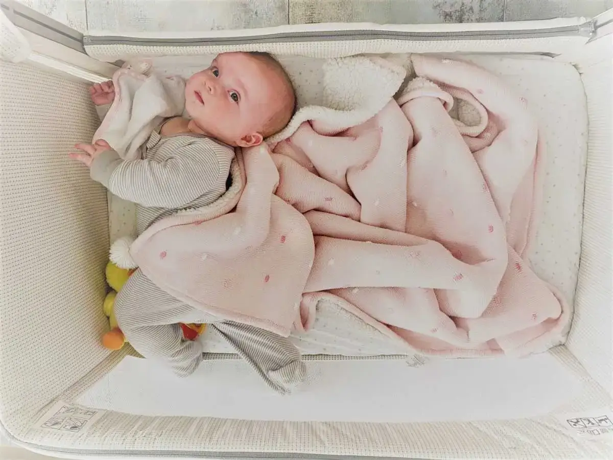 Niemowlę leży na plecach w łóżeczku, otulone różowym kocykiem. Pytanie, czy dziecko może spać na brzuchu, jest ważne dla bezpieczeństwa malucha.