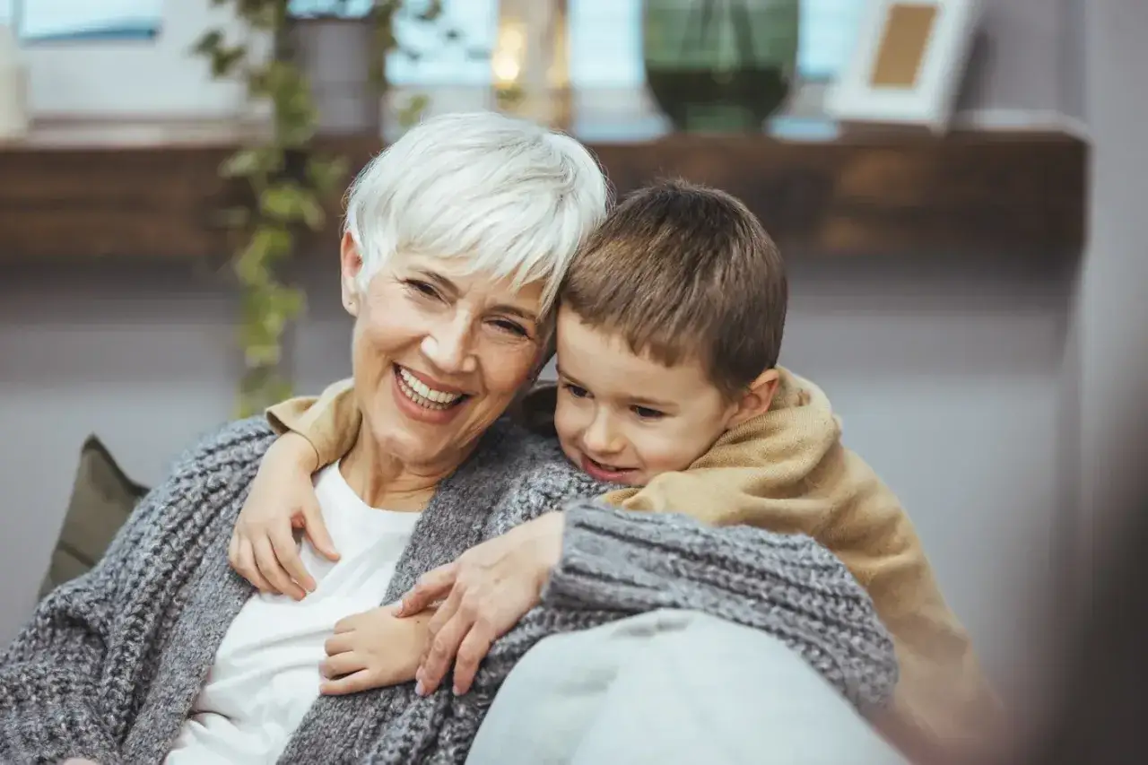 Uśmiechnięta babcia z wnukiem. Dziecko przytula się do niej, a ona cieszy się chwilą. Zastanawiamy się, ile subwencji na dziecko z orzeczeniem można uzyskać.
