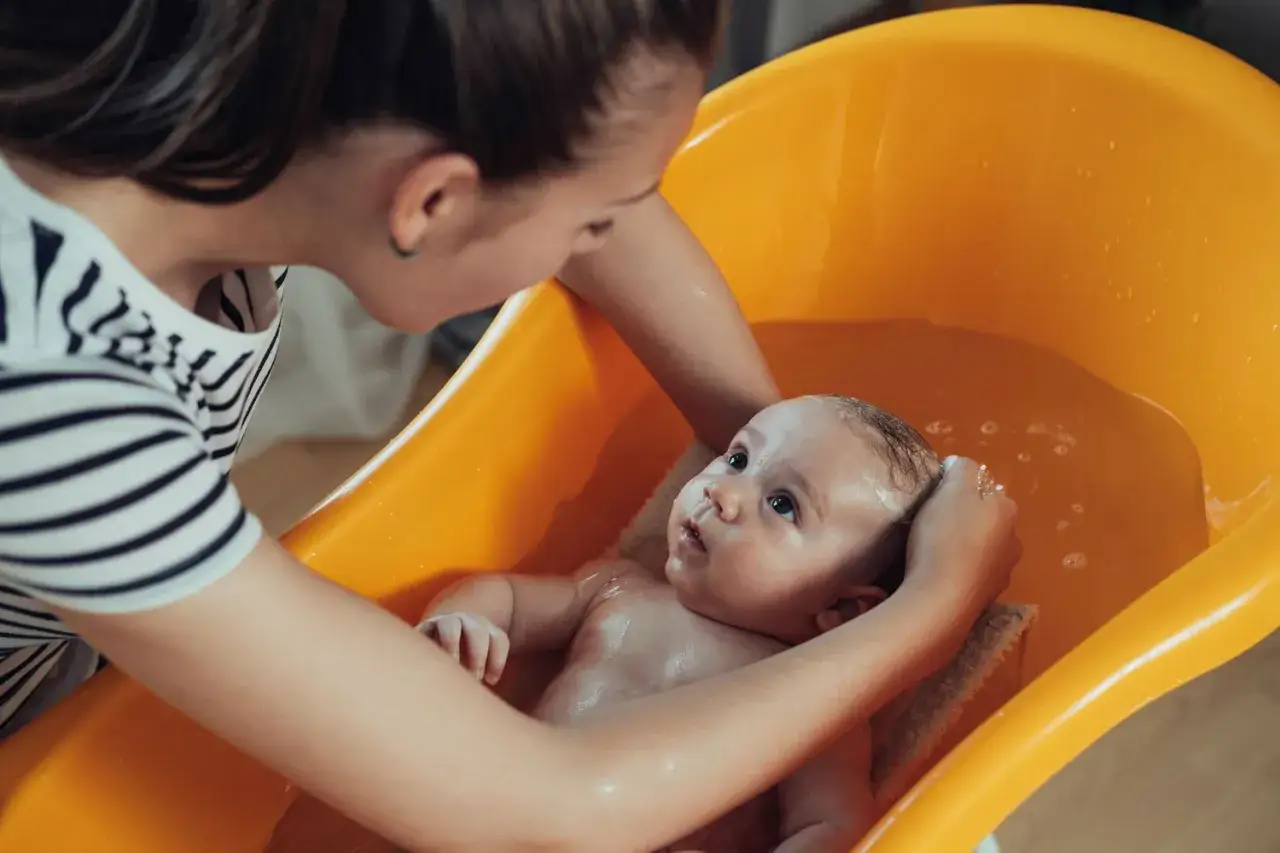 Delikatna kąpiel niemowlęcia w pomarańczowej wanience. Czy można kąpać niemowlę z gorączką? Mama delikatnie myje włoski maluszka.