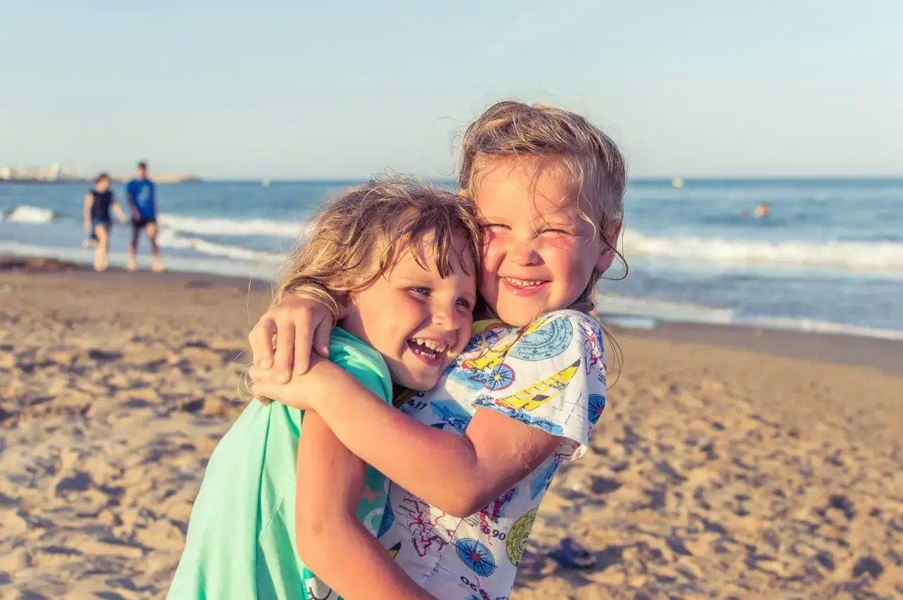 Dwie uśmiechnięte dziewczynki przytulają się na plaży. Idealne miejsce, gdzie pojechać z rocznym dzieckiem, by cieszyć się słońcem i morzem.