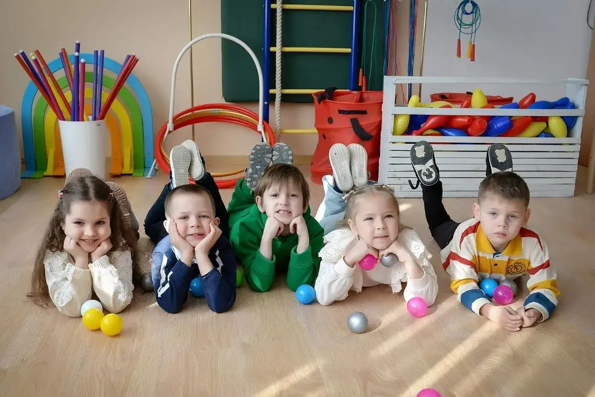 Uśmiechnięte dzieci leżą na podłodze w żłobku, gotowe na zabawę. Kolorowe piłeczki i zabawki wokół.