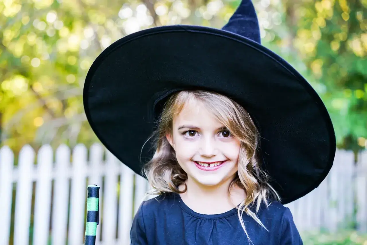 Uśmiechnięta dziewczynka w stroju czarownicy, z wielkim czarnym kapeluszem i miotłą. Idealny pomysł, jak zrobić strój czarownicy dla dziewczynki na Halloween.