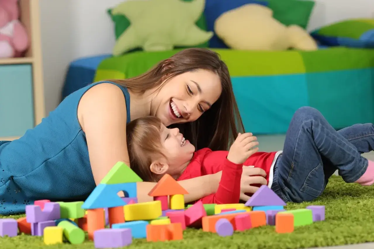 Uśmiechnięta opiekunka bawi się z dzieckiem wśród kolorowych klocków. Zastanawiasz się, ile zarabia opiekunka do dziecka za godzinę?