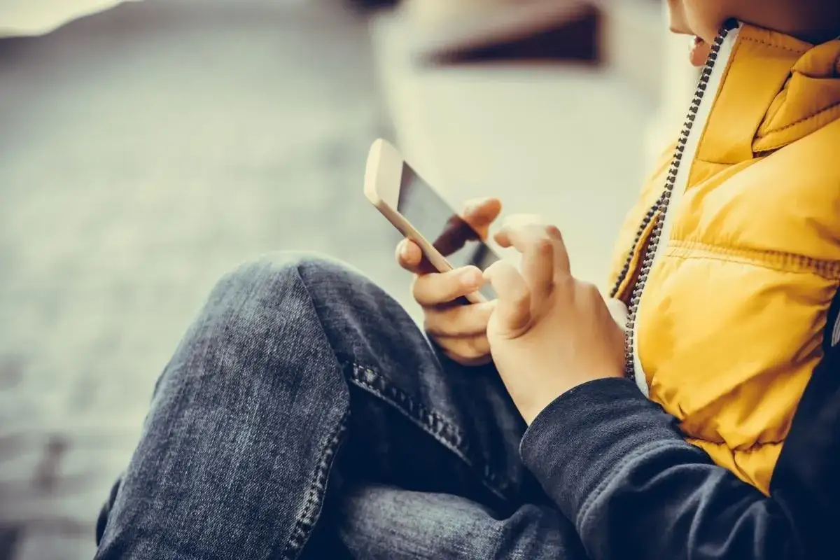 Dziecko w żółtej kamizelce i jeansach korzysta z telefonu. Rodzice szukają sposobów, jak kontrolować telefon dziecka bez jego wiedzy.