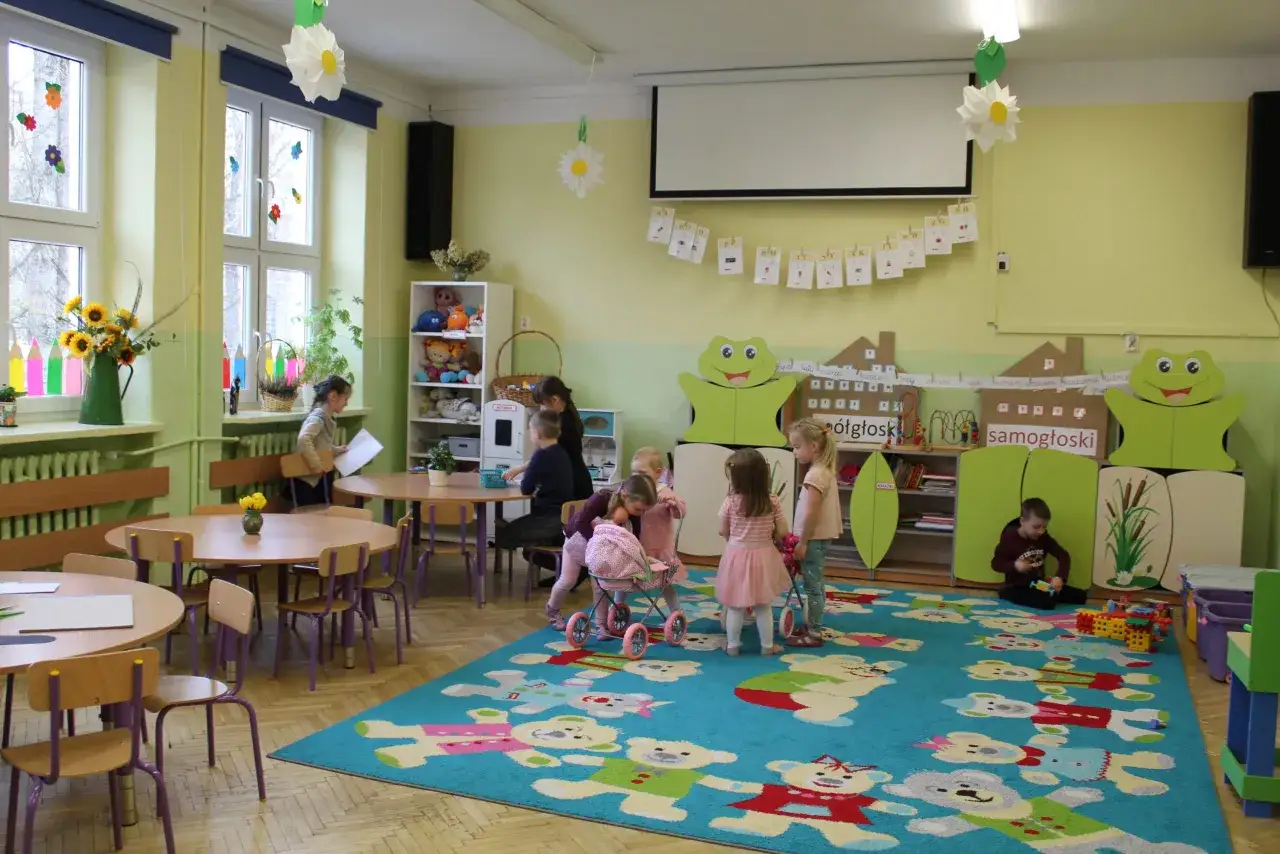 Dzieci bawią się w przedszkolu. Widać tu kolorowy dywan z misiami, stoliki, krzesła i zabawki. Wiele dzieci uczęszcza do przedszkola, by rozwijać się i bawić.