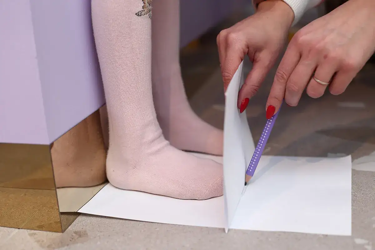 Dziecko stoi na kartce papieru, a dorosły obrysowuje jego stopę, by dobrać odpowiedni, ile większy but dla dziecka.