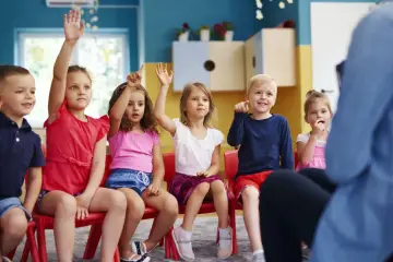 Dzieci na zajęciach w przedszkolu, z podniesionymi rękami. Widać, ile lat dziecko chodzi do przedszkola, bo są w różnym wieku.