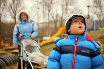 Chłopiec w niebieskiej kurtce na placu zabaw, obok wózek i kobieta. Może to być moment przed tym, jak dowiedzieć się, jak wypisać dziecko z przedszkola.